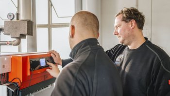 Ein Techniker justiert ein Bedienfeld an einer Heizungsanlage, während ein Kollege aufmerksam zuschaut. 