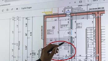 Eine Hand zeigt mit einem Stift auf einen detaillierten Bauplan, der auf einem Bildschirm angezeigt wird.