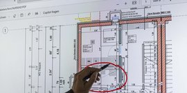 Eine Hand zeigt mit einem Stift auf einen detaillierten Bauplan, der auf einem Bildschirm angezeigt wird.