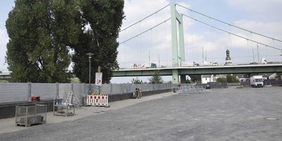 Eine grüne Brücke schwebt über einen leeren Platz, umgeben von Bäumen und Bauzäunen, während ein Lkw vorbeifährt.