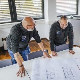 In einem hellen Raum analysieren zwei Männer gemeinsam technische Zeichnungen, während sie sich intensiv austauschen.