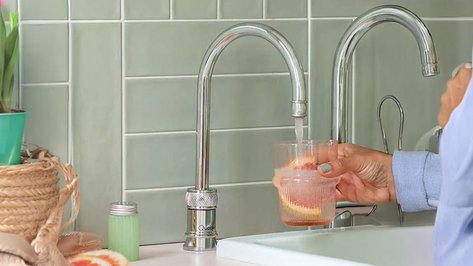 Eine Hand hält ein Glas mit Wasser und einer Orangenscheibe, während Wasser aus einem modernen Wasserhahn fließt.